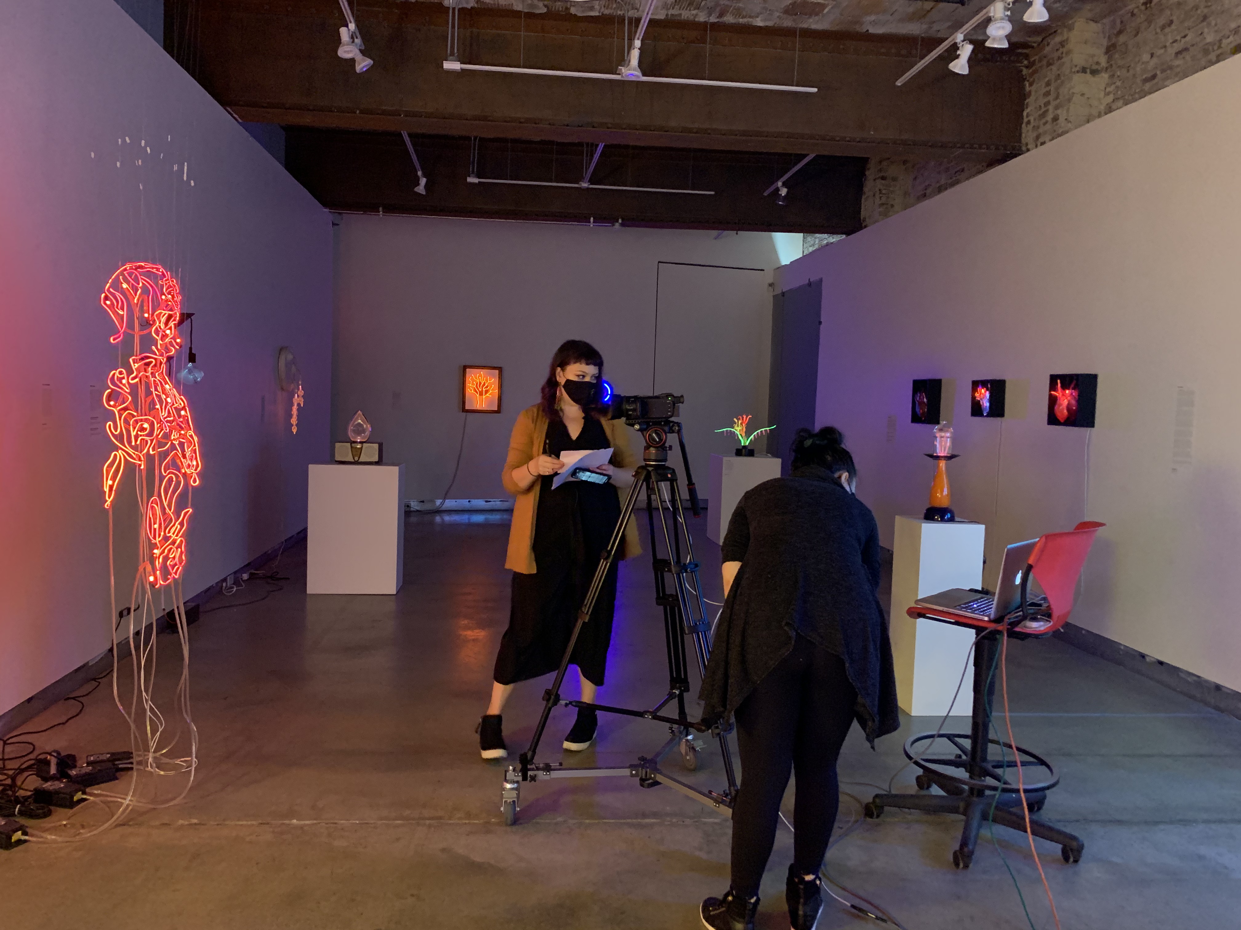 An image of my supervisor, Valerie Bundy, standing before a neon artwork in the Pittsburgh Glass Center's gallery. There is also a camera and laptop in front of her in order to communicate with the class via Zoom. Another employee stands behind the camera to help zoom in and out.