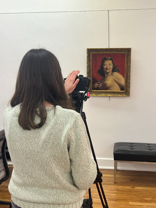 A women stands behind a camera which is pointed at a hanging framed painting of a topless woman