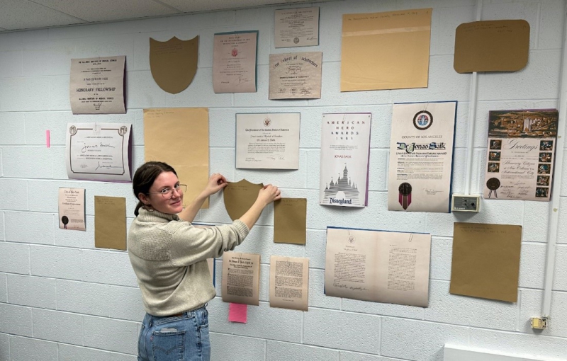 A woman stand in front of a cinderblock wall planning the layout of images and artifacts for an exhibit