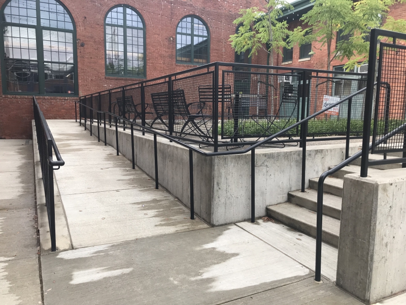 Two concrete paths both equipped with railings, one on the right with six steps of stairs, one on the left with a ramp, to access a raised concrete platform with chairs, a grassy area, trees. The platform is surrounded by a red brick building, and the single door directly parallel to 43rd street is the entrance for Associated Artists of Pittsburgh's Lawrenceville gallery space.
