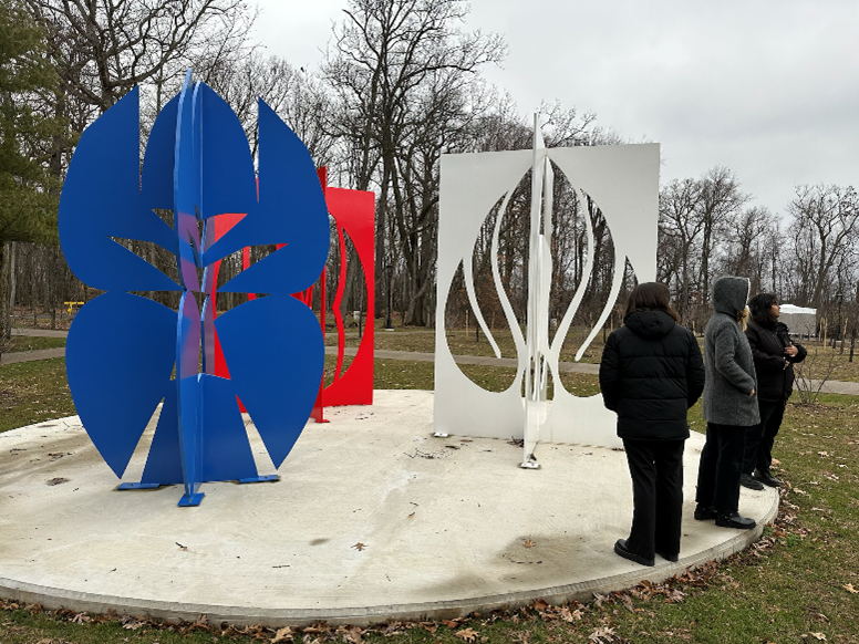Three metal sculptures in white, red and blue, stand on a concrete dais. Three people stand looking at the art