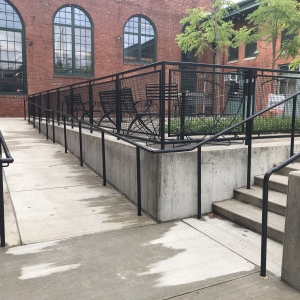 Two concrete paths both equipped with railings, one on the right with six steps of stairs, one on the left with a ramp, to access a raised concrete platform with chairs, a grassy area, trees. The platform is surrounded by a red brick building, and the single door directly parallel to 43rd street is the entrance for Associated Artists of Pittsburgh's Lawrenceville gallery space.